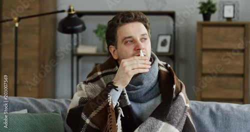 Caucasian young unhealthy caucasian man in the warm scarf clogging up his nose with medical drops as he having a bad cold. At home. Indoors