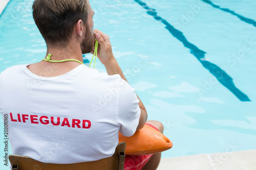 Lifeguard sitting on chair and blowing whistle at poolside photo
