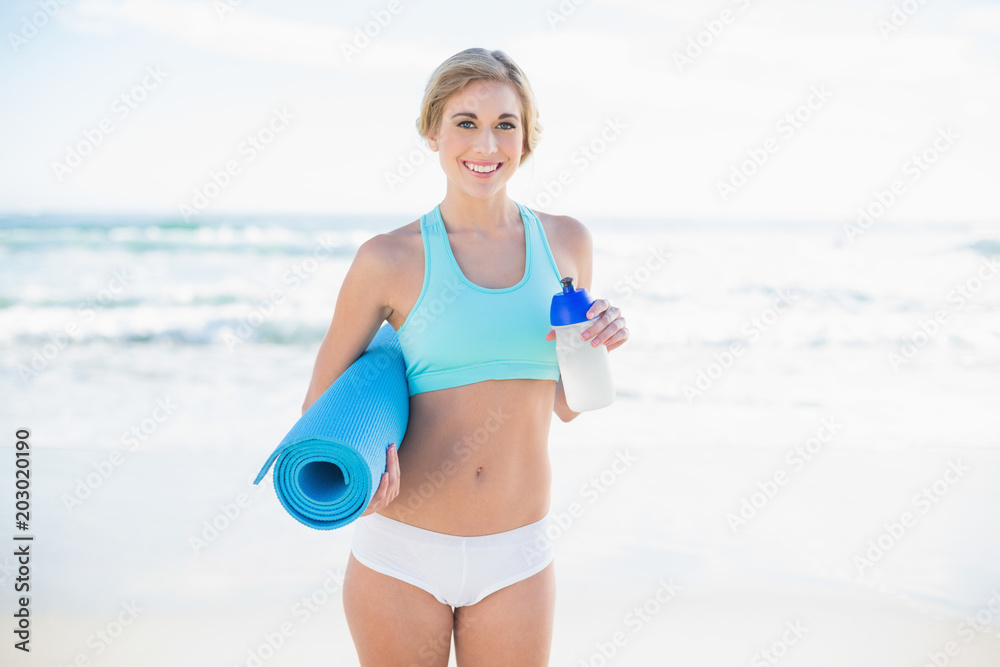 Smiling blonde woman in sportswear holding a bottle and an exercise mat Smiling blonde woman in sportswear holding a bottle and an exercise mat