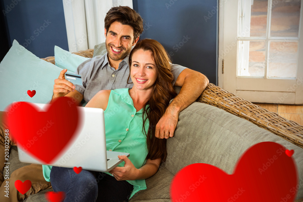 Hearts against young couple doing online shopping while sitting on sofa 3d