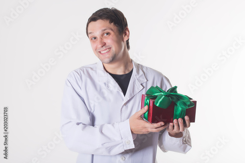 a man in a white robe holds a gift box. smiling doctor with a red box with a green bow. Handsome man looking at camera on white background