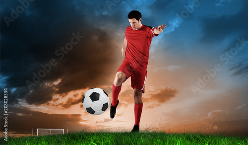Football player in red kicking against green grass under blue and orange sky