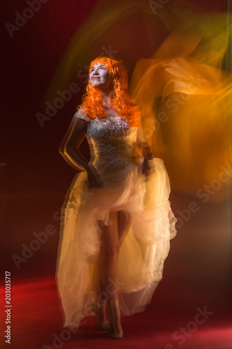 Fototapeta Naklejka Na Ścianę i Meble -  Old female actress with orange hair dancing in white dress with light show with yellow, orange and green lights around