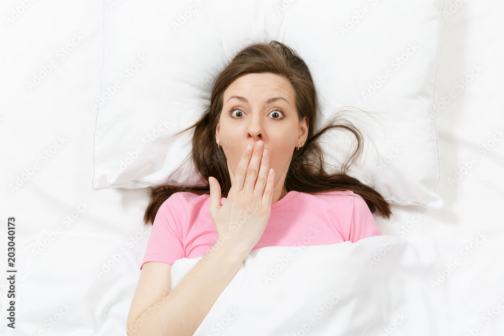 Top view of happy brunette young woman lying in bed with white sheet, pillow, blanket. Female spending time in room, cover mouth with hand. Rest, relax, good mood concept. Copy space for advertisement Top view of happy brunette young woman lying in bed with white sheet, pillow, blanket. Female spending time in room, cover mouth with hand. Rest, relax, good mood concept. Copy space for advertisement