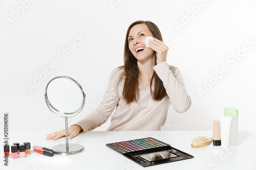 Attractive fun woman wipes her skin face with facial sponges, sits at table applying makeup with set facial decorative cosmetics isolated on white background. Beauty female fashion lifestyle concept.