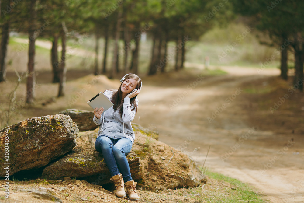 Young relaxing smiling woman in casual clothes sitting on stone listening music with headphones and tablet pc computer in city park or forest on green background. Student lifestyle, leisure concept.