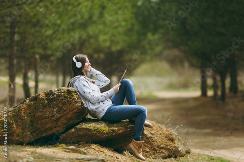 Young relaxing woman in casual clothes sitting on stone listening music with headphones and tablet pc computer in city park or forest on green blurred background. Student lifestyle, leisure concept.