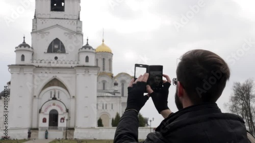 Young brunette black-haired man with glasses standing on an observation deck on the hill in the park of Pushkin in Vladimir, taking pictures and selfie near Uspenskiy Sobor Cathedral. photo