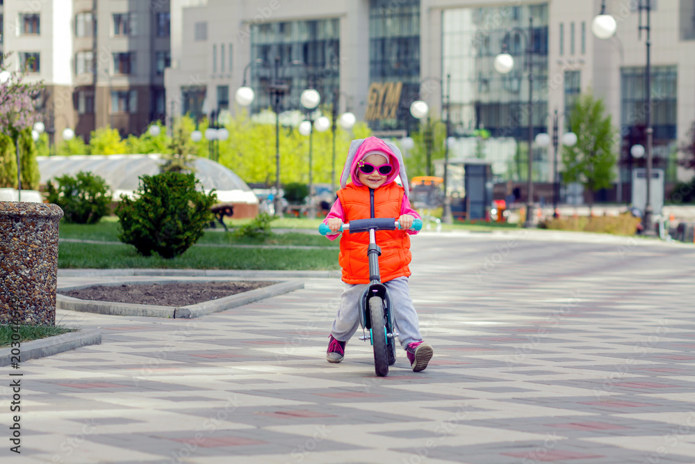 Active blond kid driving run bike the street alley of the city. Funny casual clothing vest jacket, sunglass and hood with bunny ears. Balance bike concept. Active blond kid driving run bike the street alley of the city. Funny casual clothing vest jacket, sunglass and hood with bunny ears. Balance bike concept.