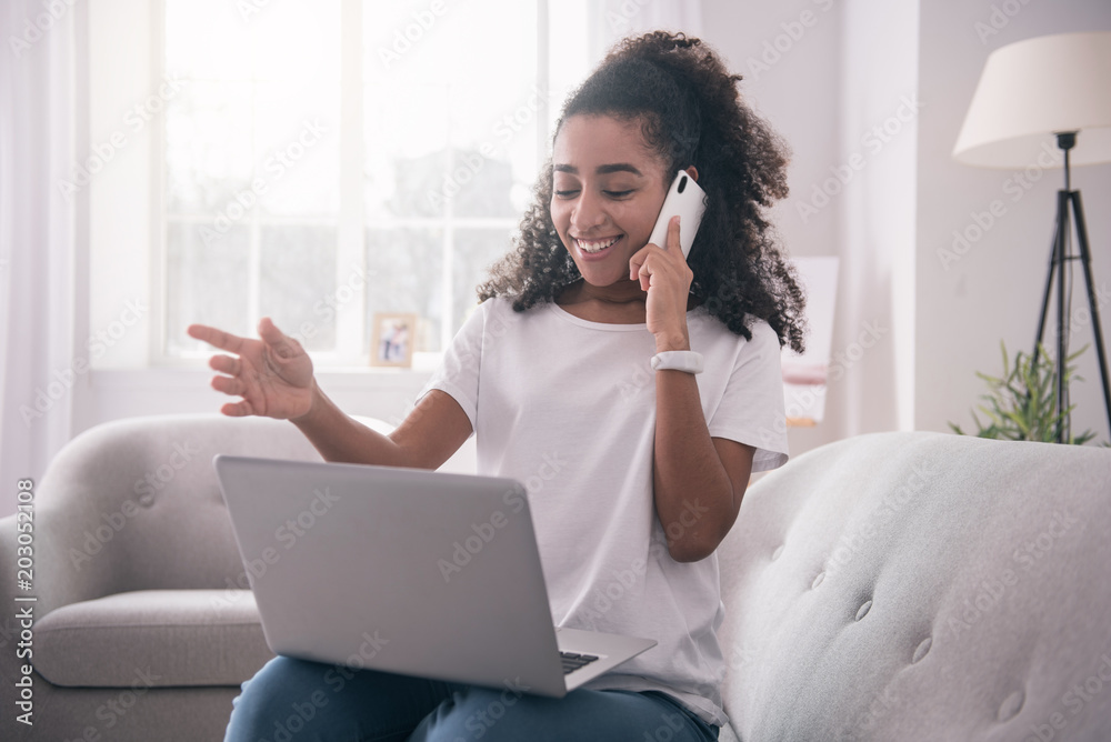 Pleasant interaction. Joyful young woman looking at the laptop screen while enjoying her phone conversation with her university friend Pleasant interaction. Joyful young woman looking at the laptop screen while enjoying her phone conversation with her university friend