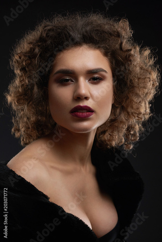 Sexy curly woman posing in the studio and looking at camera. Looking beatiful and passionate. Wearing light day make up and saturated violet lipstick on plump lips. Black background.