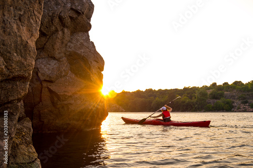 Young Woman Paddling the Red Kayak on Beautiful River or Lake near High Rocks at Sunset © Maksym Protsenko