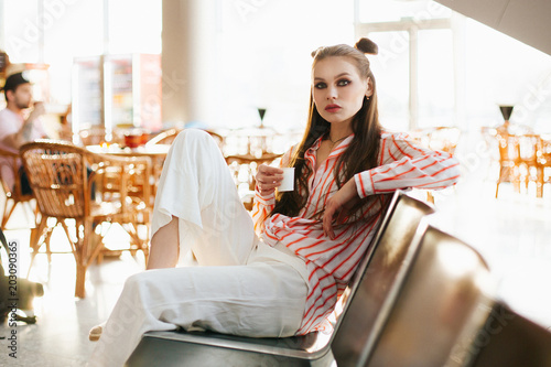 Young fashion model sits with a cup of coffee on the chairs in the airport