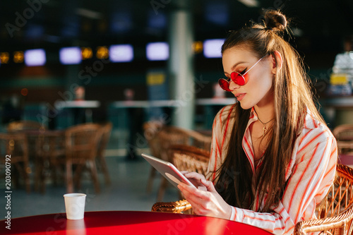 Pretty fashion model in red sunglasses works with her tablet sitting at the cafe in the rays of sunset