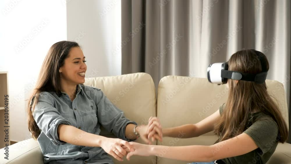 Two girlfriends on the couch in the living room having a lot of fun. The youngest is using a VR headset while the oldest is looking at her and laugh. Static shot Two girlfriends on the couch in the living room having a lot of fun. The youngest is using a VR headset while the oldest is looking at her and laugh. Static shot