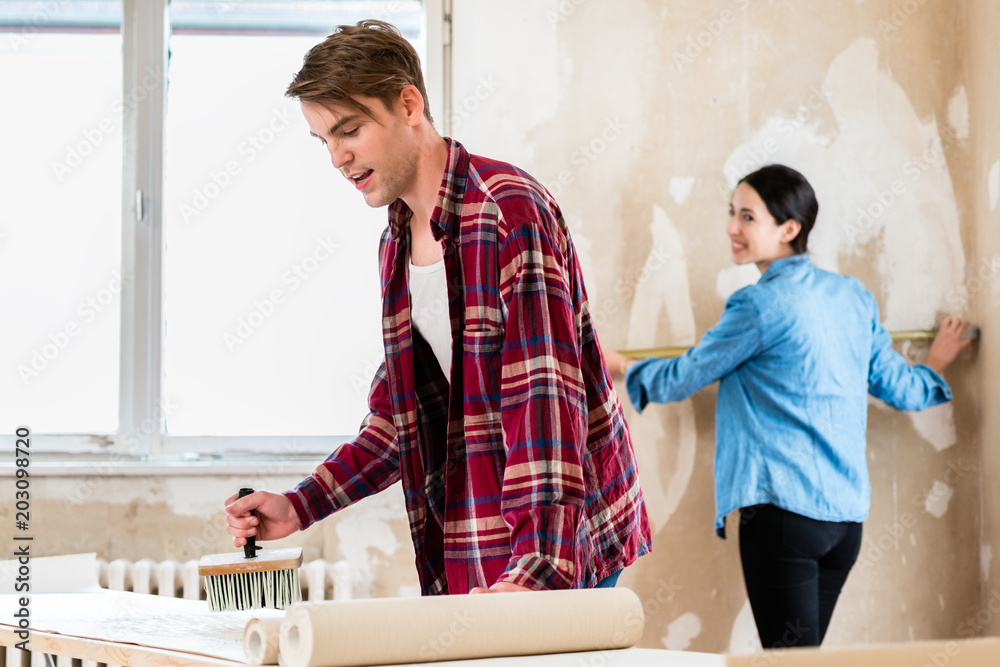 Obraz premium Young man applying adhesive to the back of the wallpaper sheet while his girlfriend is measuring the prepared wall in their new home