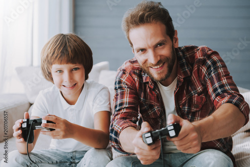 Cheerful father with game controller plays video game with small son sitting on couch.