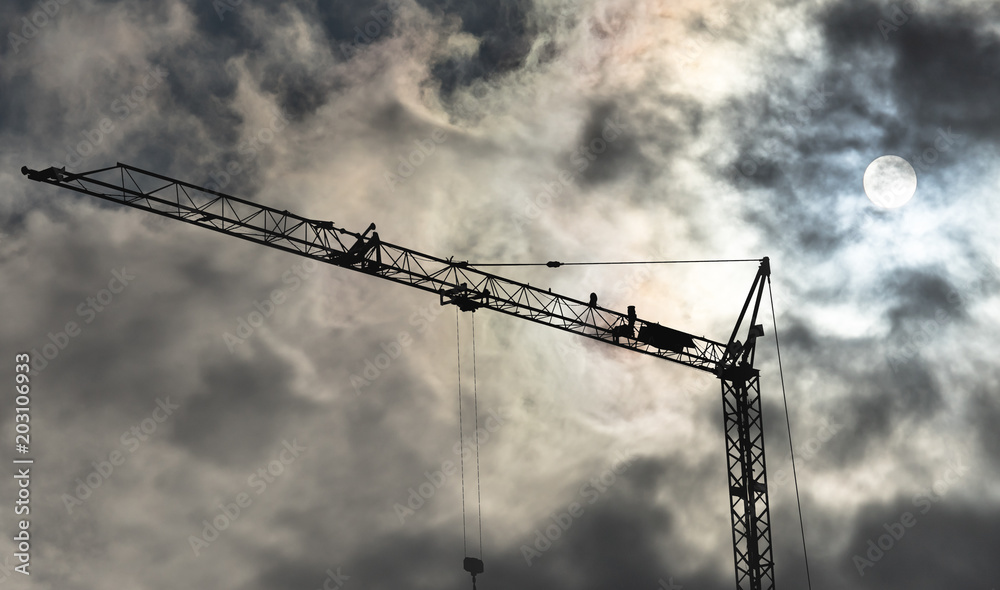 High black construction crane in front of a dramatic threatening sky with translucent sun behind the clouds High black construction crane in front of a dramatic threatening sky with translucent sun behind the clouds