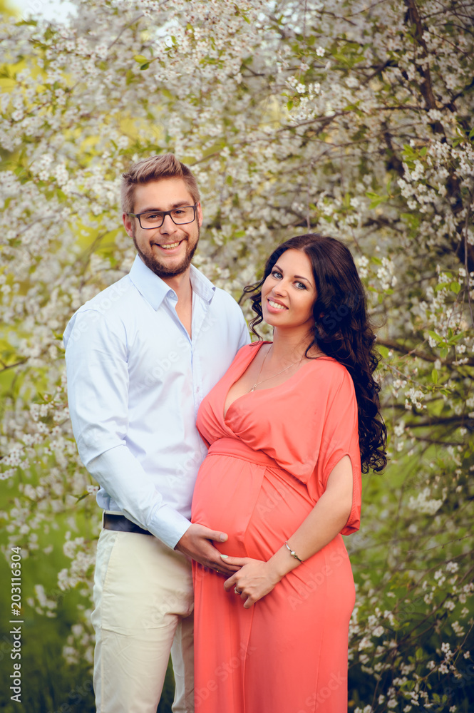 Romantic portrait of young smiling happy couple of lovely future parents during sunset on nature apple tree background in the city park. Pregnancy pregnant future mother photoshoot. Motherhood photo Romantic portrait of young smiling happy couple of lovely future parents during sunset on nature apple tree background in the city park. Pregnancy pregnant future mother photoshoot. Motherhood photo