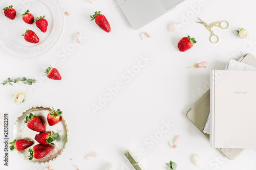 Frame of feminine modern home office desk with laptop, notebook, lipstick, fresh raw strawberries and rose flower buds on white background. Flat lay, top view.
