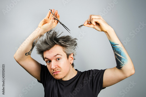 Unshorn and unshaven young guy with piercings on his face, shears her hair and looks in camera, on gray background. photo