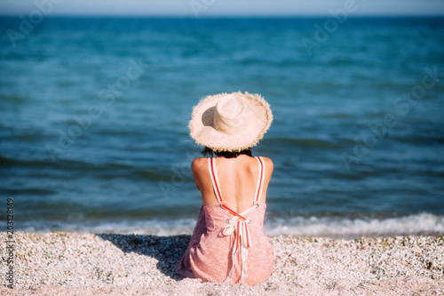 Fotografía Young girl in a hat sits on the beach at sunset, happily travels and enjoys the