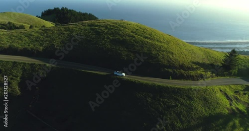 Aerial view of car driving down country road through rural rolling hills with ocean in background at sunset