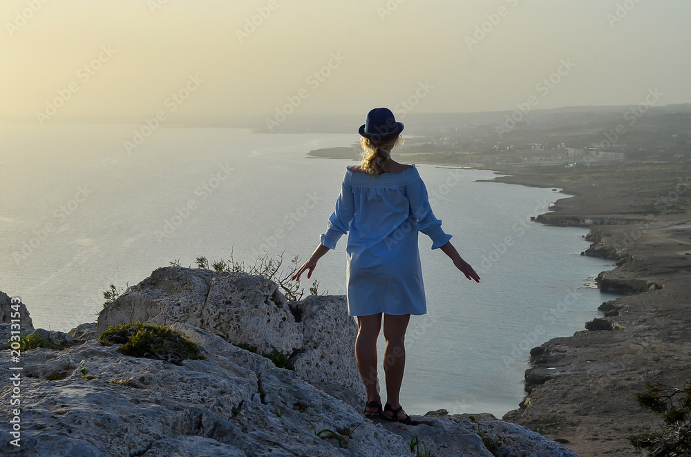 Woman traveler standing near sea on top of cliff in summer mountains and enjoying view of nature. Girl in blue dress and straw hat on sunset. Cape Greco, Cyprus, Mediterranean Sea