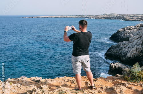 Traveler man taking photo with mobile phone standing near sea. Hiking man standing on top of cliff in Cape Greco, Cyprus, Mediterranean Sea. Travel, vacation concept. Copyplace, place for text photo