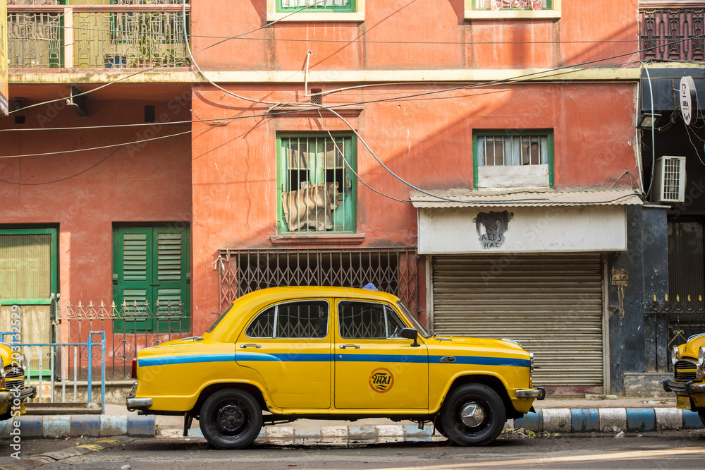 An Ambassador yellow cab taxi is parked in the street under a red-colored house in Kolcata, India