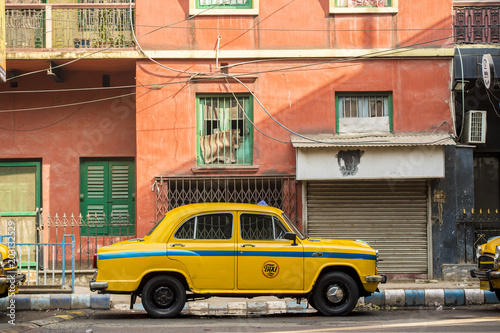An Ambassador yellow cab taxi is parked in the street under a red-colored house in Kolcata, India © Travel Wild