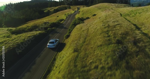 Aerial view of car driving down country road through rural rolling hills at sunset
