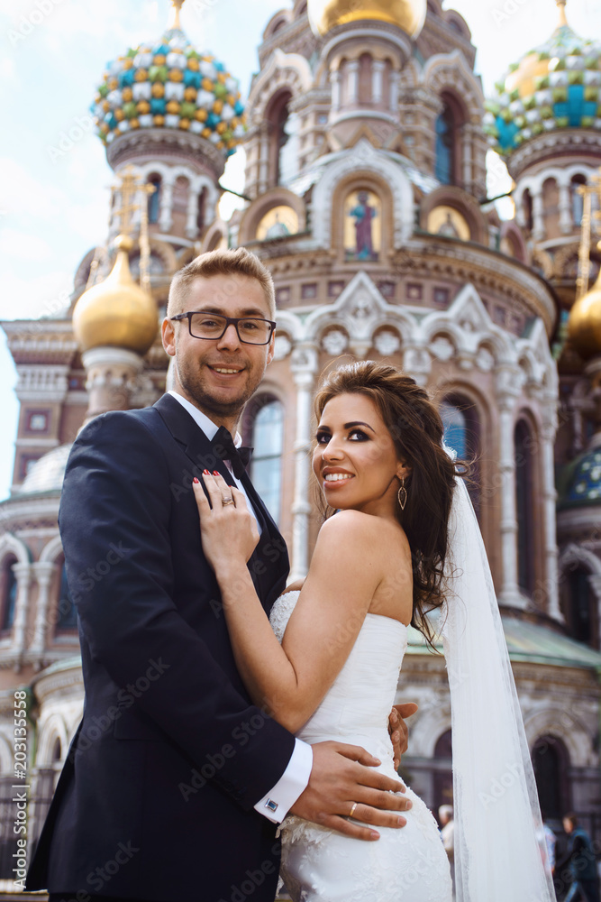 Newlywed couple bride and groom photoshoot after wedding ceremony in Saint-Petersburg. Church of the Savior on blood. White wedding dress with long veil and classical black suit or smoking with bowtie