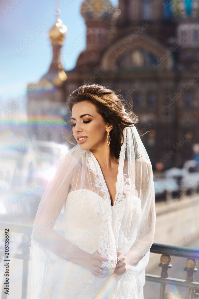 Naklejka premium Close up portrait of beautiful young caucasian brunette woman bride in white wedding dress over Saint-Petersburg on blurred background. Smiling girl in wedding dress