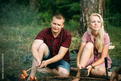 Beautiful couple in love sitting by campfire. Romantic date in nature. Blond girl in nice retro dress drinking tea while her boyfriend cooks food