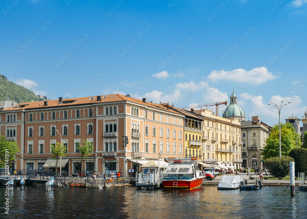 View of the city of Como, including boats on the marina and traditional colours buildings on the waterfront of Lake Como.