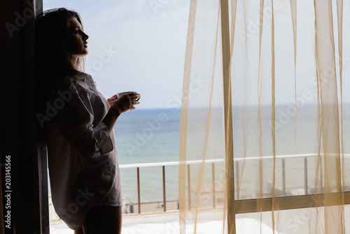 Cheerful beautiful woman enjoying healthy relax lifestyle drinking hot beverage looking out window having positive day and bright sunny oceanfront outdoors background. Travel relaxation destination