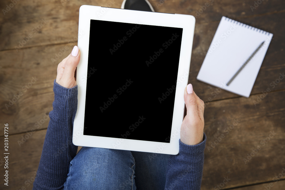 The girl sits on a wooden floor and holds a tablet with a blank screen in her hands. Notebook on the floor. Top view. The girl sits on a wooden floor and holds a tablet with a blank screen in her hands. Notebook on the floor. Top view.
