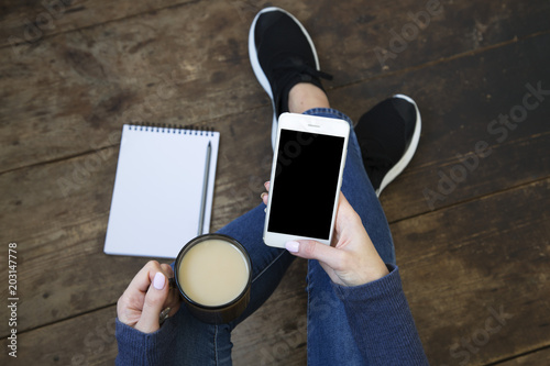 The girl sits on a wooden floor and holds a smartphone with a blank screen and latte in her hands. Notebook on the floor. Top view.