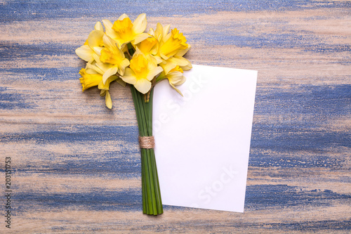 Fototapeta Naklejka Na Ścianę i Meble -  A bouquet of daffodils and a sheet of white paper on a wooden background. Spring yellow flowers. Paper with space for text. Blue peeling paint. Flat lay design, top view.