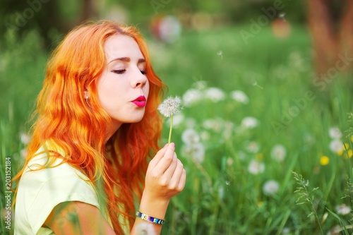 Young spring fashion redhead woman blowing dandelion in spring garden. Springtime, summertime. Trendy girl in summer landscape background. Allergic to pollen of flowers. Spring allergy
