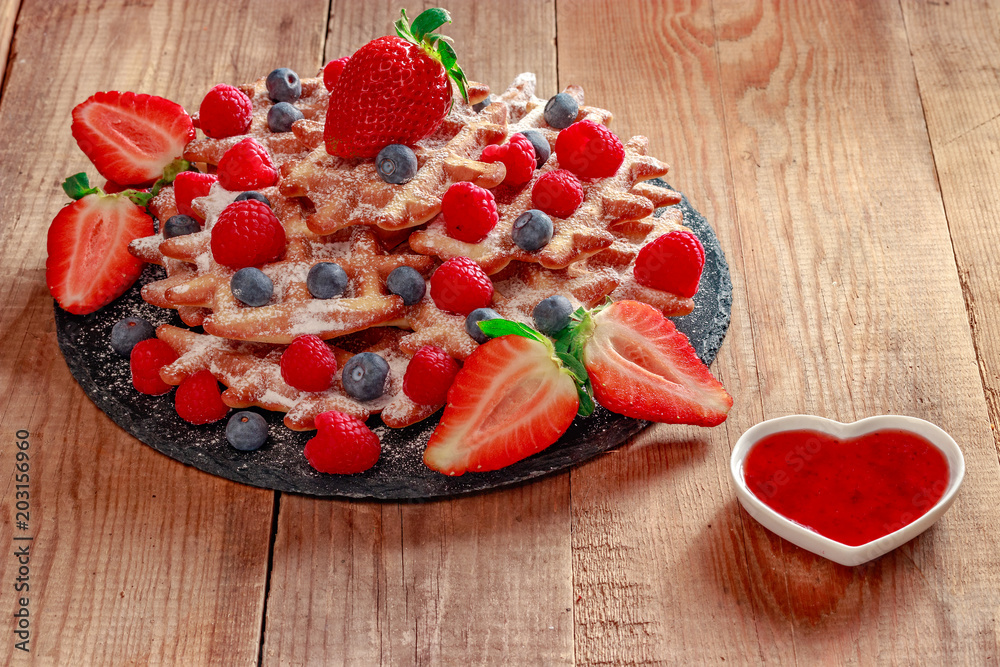 On a stone black dish waffles sprinkled with white powdered sugar. Viennese waffles are decorated with sliced strawberries, blueberries and raspberries. On the boards is a heart-shaped saucer with jam