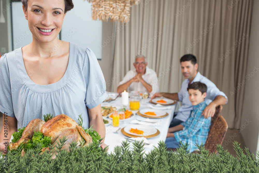 Woman holding chicken roast with family at dining table against green fir branches