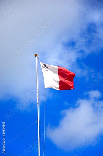 Maltese flag unfurled against a blue sky with fluffy clouds, Malta.