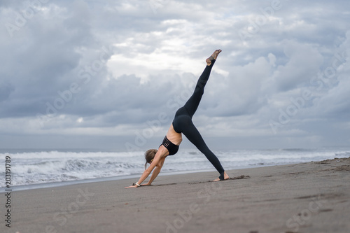 sporty young woman practicing yoga in One Legged Downward-Facing Dog pose (Eka Pada Adho Mukha Svanasana) on seashore