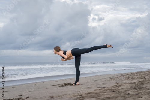 sporty young woman practicing yoga in Warrior III pose (Virabhadrasana III) on seashore