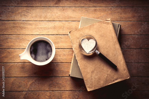 White cup with coffee and books with magnifier, heart on wooden table. High angle view