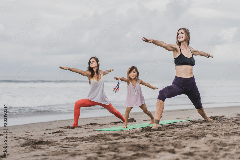 two women and little child practicing yoga on seashore on cloudy day, Warrior pose (Virabhadrasana) two women and little child practicing yoga on seashore on cloudy day, Warrior pose (Virabhadrasana)