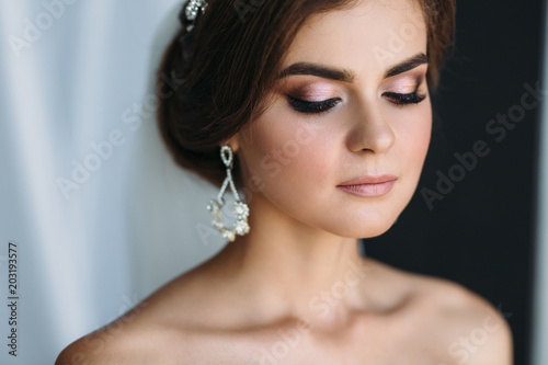 Close-up portrait of the bride with diamond earrings, wedding make-up and hairdo poses in a dark studio. Beautiful young brunette girl on black and white background. Concept of marriage, clear skin