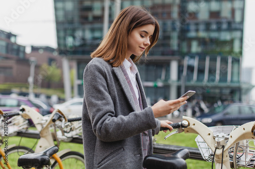 Attractive office worker using cell phone, young stylish girl using bicycle rent mobile app smiling outdoors, portrait of female manager browsing smartphone standing near bike sharing to go cycling